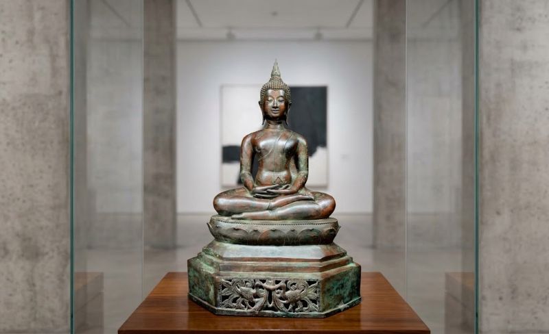 A bronze Buddha statue seated in the lotus position atop a decorated, carved pedestal, displayed inside a clear glass showcase within a modern gallery space